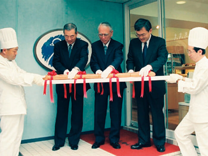 スワンベーカリー1号店（銀座）のオープンセレモニー。 左から有富慶二（当時のヤマト運輸社長）、小倉昌男初代理事長、タカキベーカリー高木社長（1998年）